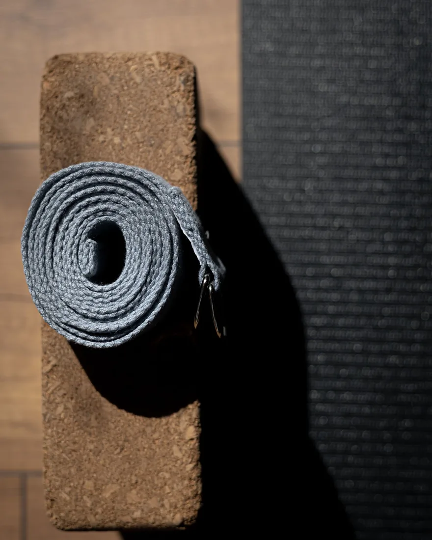 Cinturón de yoga enrollado sobre un bloque de corcho junto a una esterilla oscura en un suelo de madera, con luz rasante